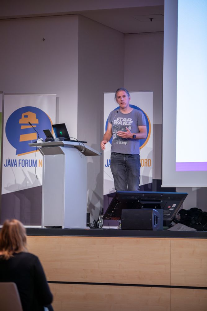 Eberhard Wolff steht auf der Bühne am Rednerpult, spricht mit ernstem Gesichtsausdruck und gestikuliert mit den Händen. Er trägt ein graues Star-Wars-T-Shirt und ein Headset. Hinter ihm sind Banner mit dem Logo des Java Forum Nord zu sehen, im Vordergrund das Publikum.