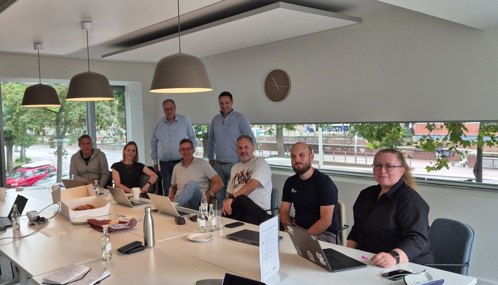 Acht Personen des SWAGLab-Teams sitzen und stehen in einem hellen Besprechungsraum mit Laptops, Getränken und Snacks auf dem Tisch, Blick in die Kamera.