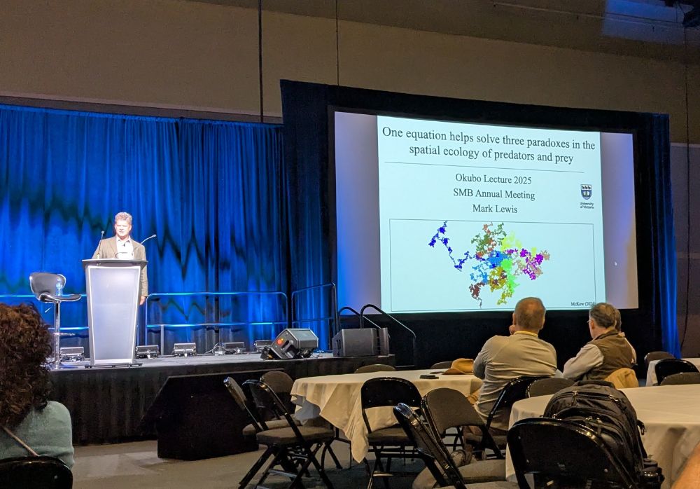 Mark is on the left of the stage at the podium. On the right is his presentation entitled: One equation helps solve three paradoxes in the spatial ecology of predators and prey.