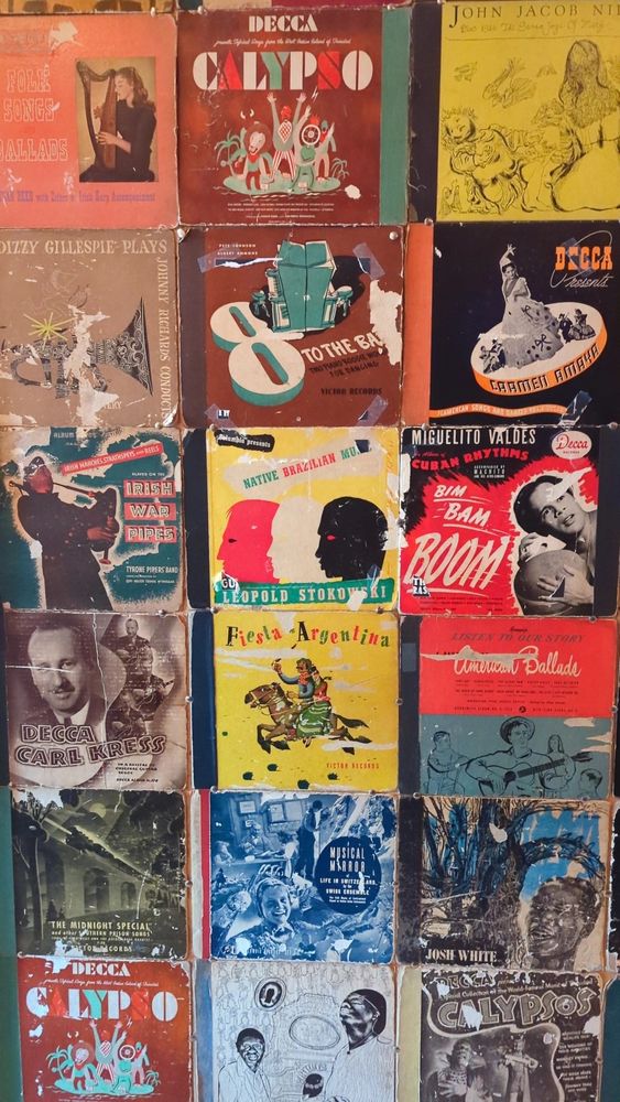 Photo of 1045 rpm vinyls hung up on a wall. Vinyls included are by artists like Dizzy Gillespie,and Decca records of Calypso and Folk songs