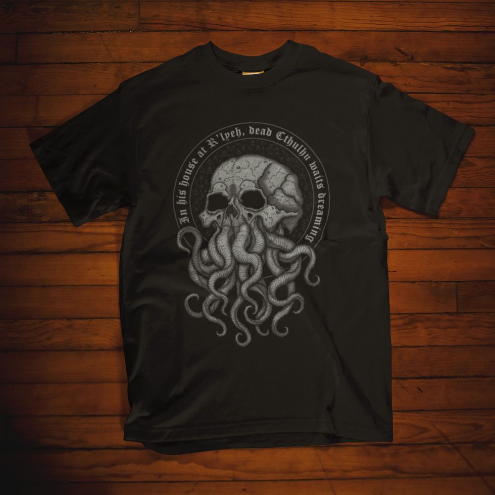 A t-shirt depicts a detailed black and white illustration depicts a skull-faced Cthulhu with tentacles in place of its mouth. The skull is rendered in a realistic style, with intricate details of bone structure and texture. The tentacles are numerous and sinuous, creating a sense of unease and dread. The image is framed by a circular border with the inscription "In his house at R'lyeh, dead Cthulhu waits dreaming."