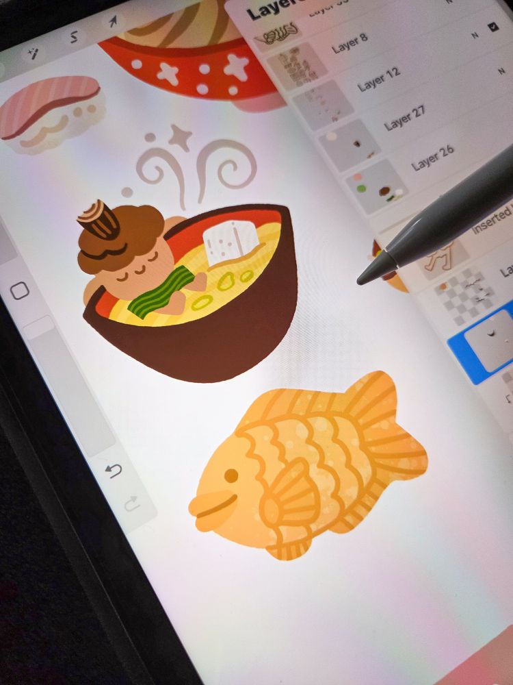 A photo of ronnie working on a Japanese Food sticker sheet. There is a drawing of an acorn relaxing in a bowl of miso soup and below, a drawing of Taiyaki 