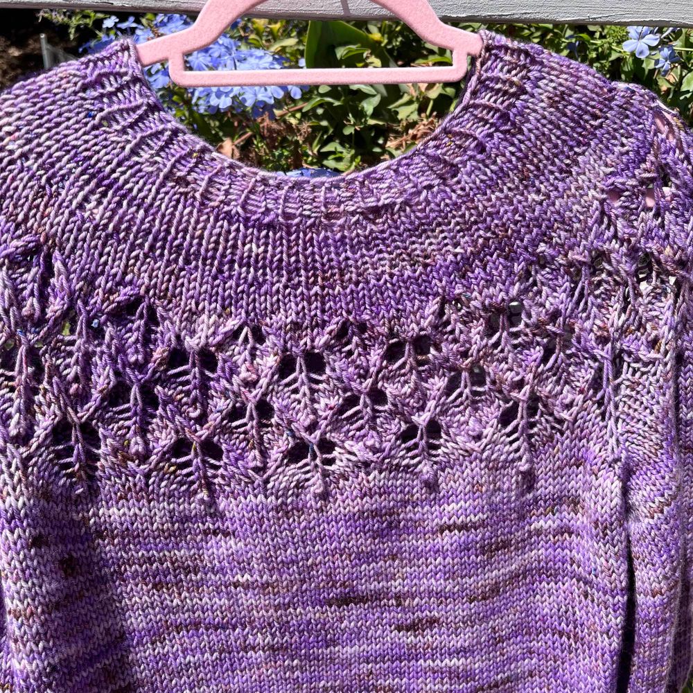 A cropped Love Note sweater with a lace yoke hangs from a white fence rail in front of a plumbago plant blooming with blue flowers.  The sweater is a tonal lavender with flecks of brown scattered throughout.