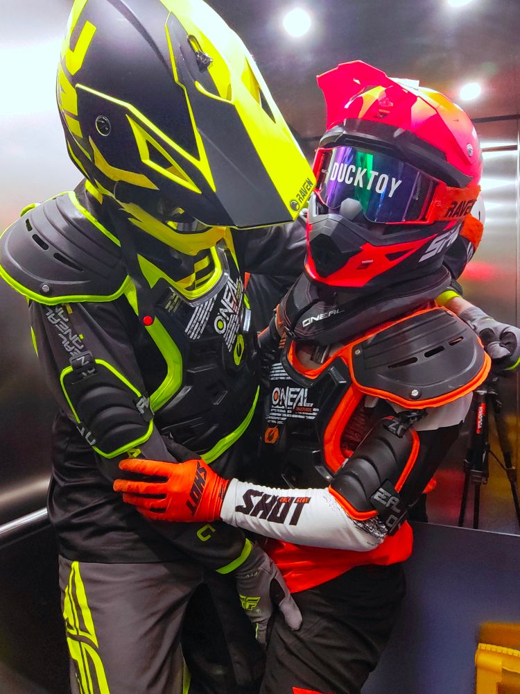 Two people in a lift/elevator in full Motocross gear, including helmets and body armour. The left person (@astra.blue) is wearing yellow gear, groping the right persons' (@ducky.ws; it/its) crotch, who is wearing orange gear with letters across its goggles that read "DUCKTOY".