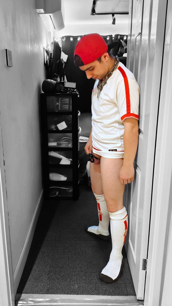A person (it/its) stood in an open doorway into a room, void of any bottom clothing to show off their cock locked in a chastity cage. It's also wearing white/red football socks and football jersey, a red flat-billed cap worn backwards, and a locked metal chain collar.