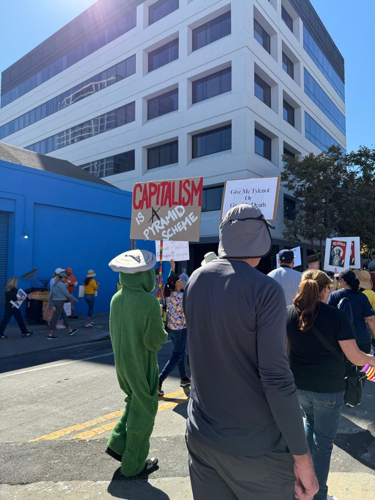 A person dressed as Oscar the Grouch holding a sign that reads “ Capitalism is a pyramid scheme”