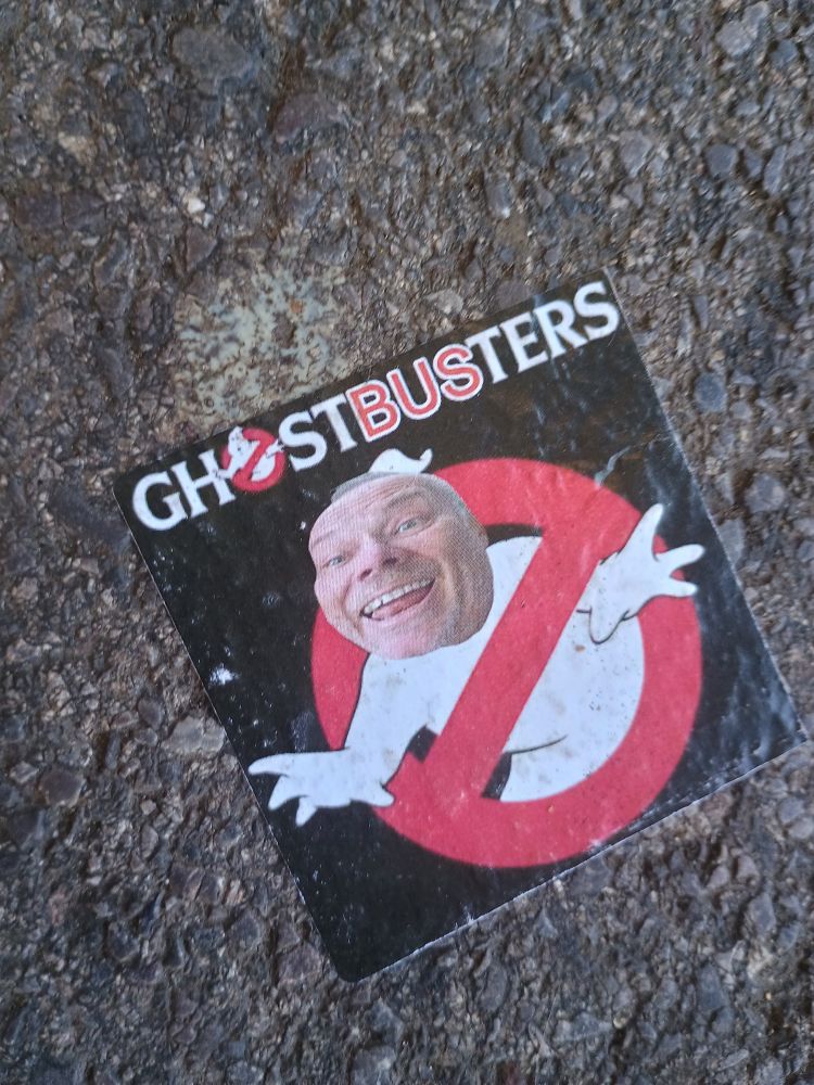 Sticker found on asphalt showing a smiling middle aged white guy's face superimposed on the Ghostbusters logo, with the letters BUS highlighted in red