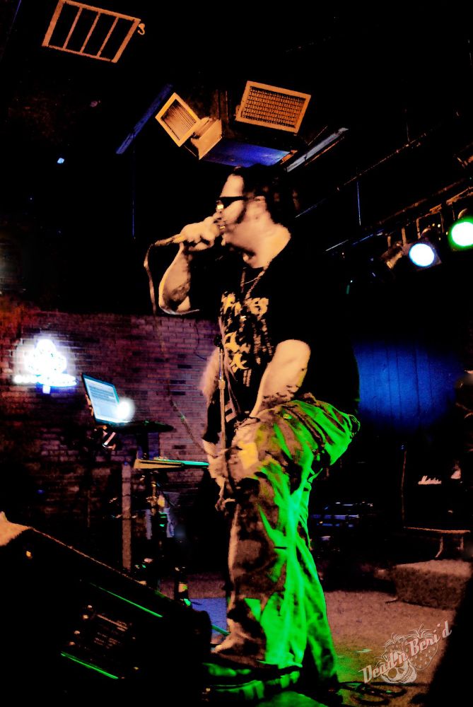 A tattooed singer with pork chop sideburns and a pomp haircut  profile side view in baggy camo pants, newrock boots.  In the foreground is a stage monitor, in the background is ankeyboard stand with a synthesizer and a laptop.  There is a brickwall stage right, black curtains upstage Nd some par cans are suspended with green light