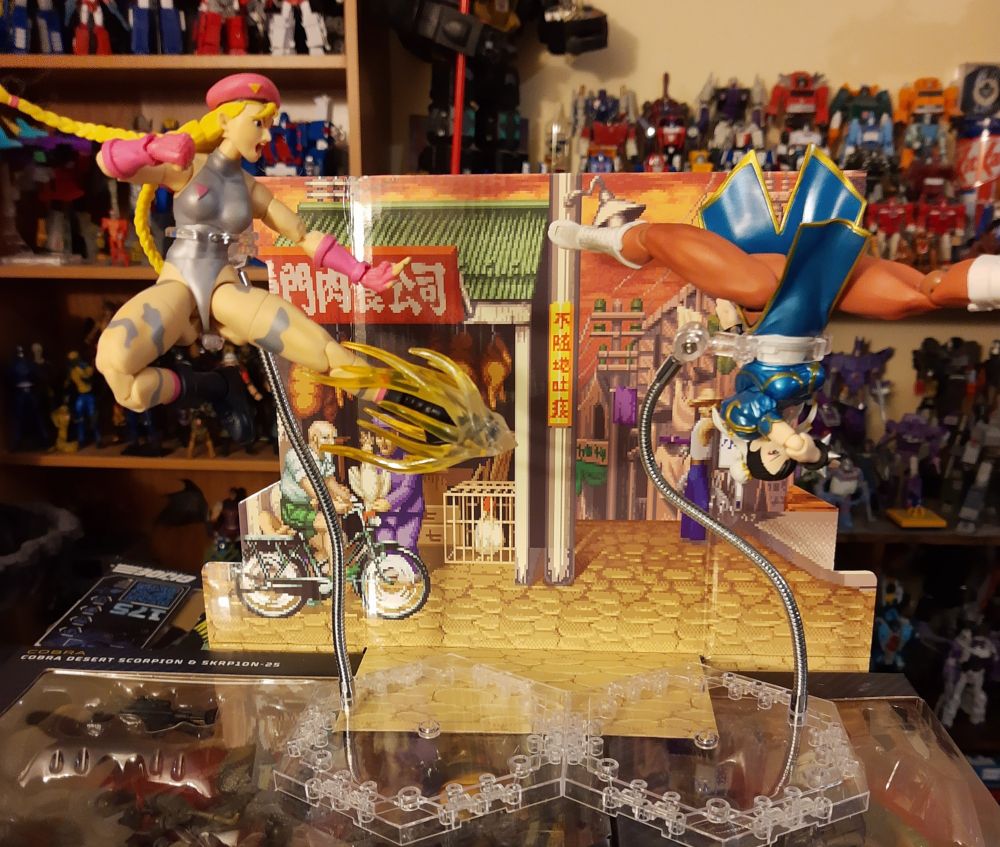 A shot focused on two Street Fighter 2 action figures, Cammy on the left and Chun-Li on the right, both on flight stands in front of a cardboard scene of the Chinese street stage.
Cammy is doing a single leg jumping kick to the right with a special effect accessory on her forward foot.
Chun-Li is upside down and doing the spits, it is implied she is spinning in a corkscrew manner.
