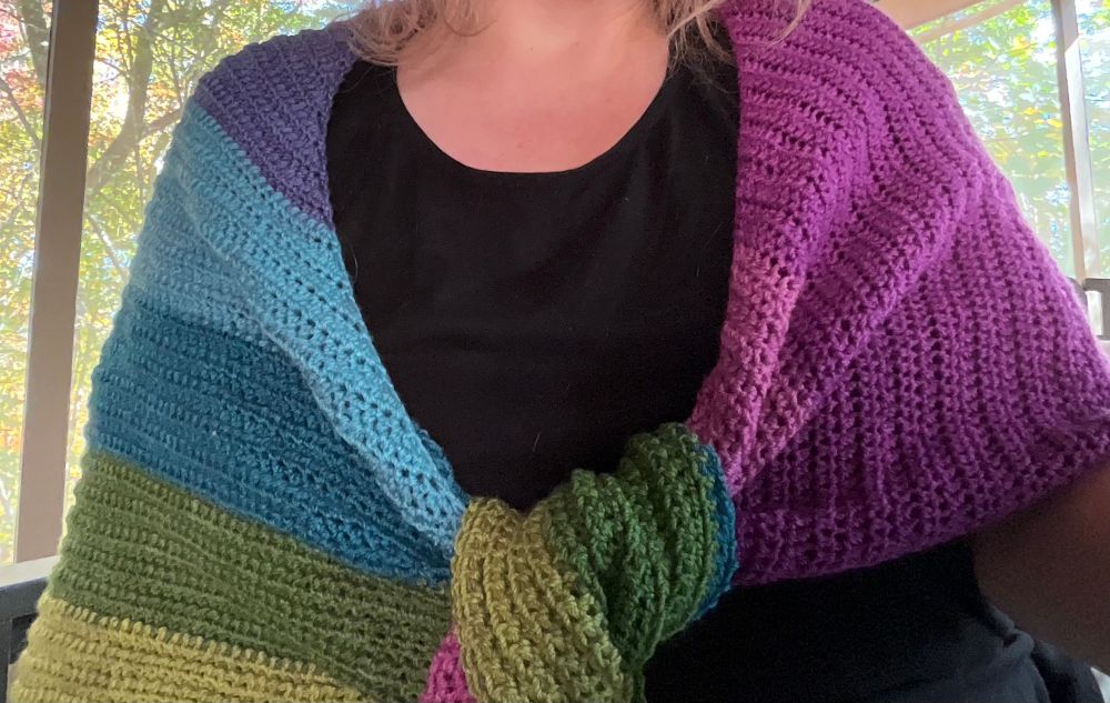 Torso of a person wearing a triangle shawl with the ends knotted in front over a simple black tee. The shawl has wide stripes in six colors ranging from from red-violet to blue to yellow-green.