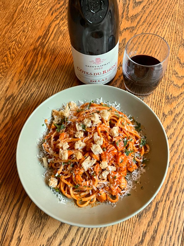Stoneware bowl of linguini with a red sauce with fish chunks, garnished with chopped parsley and tiny torn sourdough croutons. Bottle of Rhone red blend and a stemless wine glass of same.