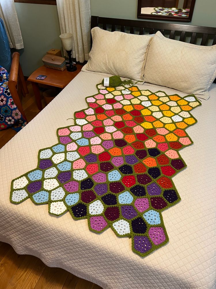 A Cairo tiling crocheted throw laid out on a bed, with motifs in a medley of colors found in flowers and joined in a medium-dark leaf green