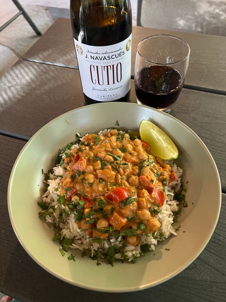 Stoneware bowl of rice and a light red-orange sauce with red bell pepper, fresh roughly chopped spinach, and chickpeas, garnished with a lime wedge. Wine pairing: Spanish Garnacha-Cariñena blend.