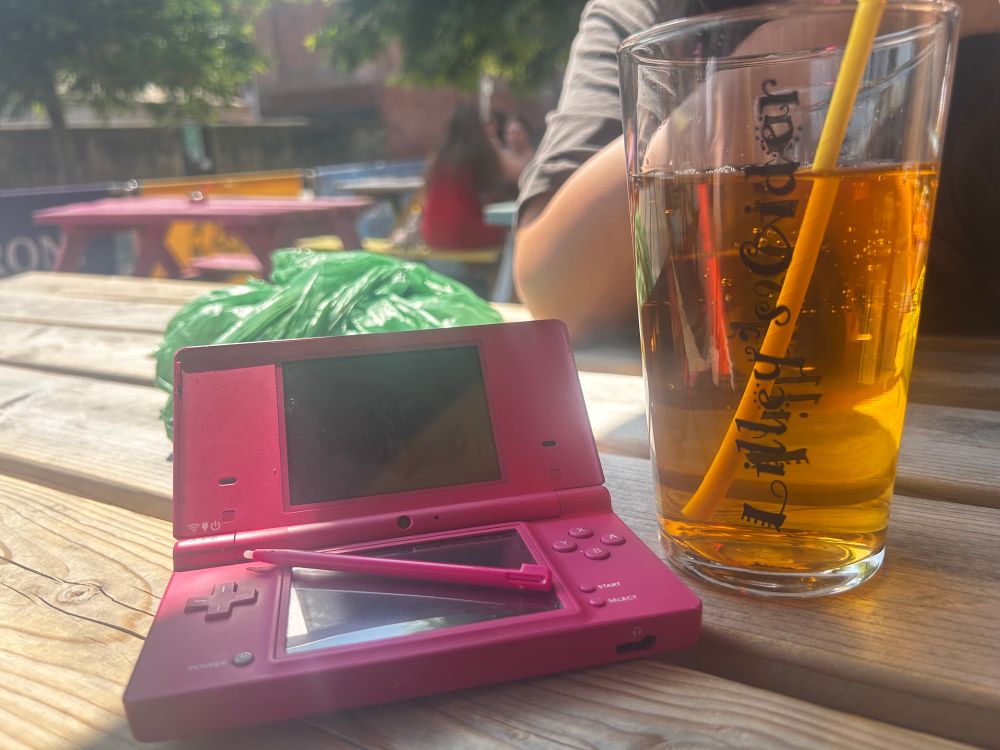 a nintendo dsi on a table in the shade next to a glass of lileys mango 