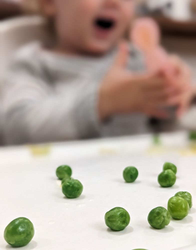A close up photo of peas on a baby's feeding tray, with said baby in soft focus in the background 