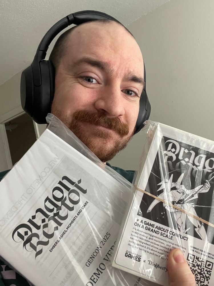 Quinn, smiling, holding the Gencon Demo Version of Dragon Reactor and some similarly themed stickers. 