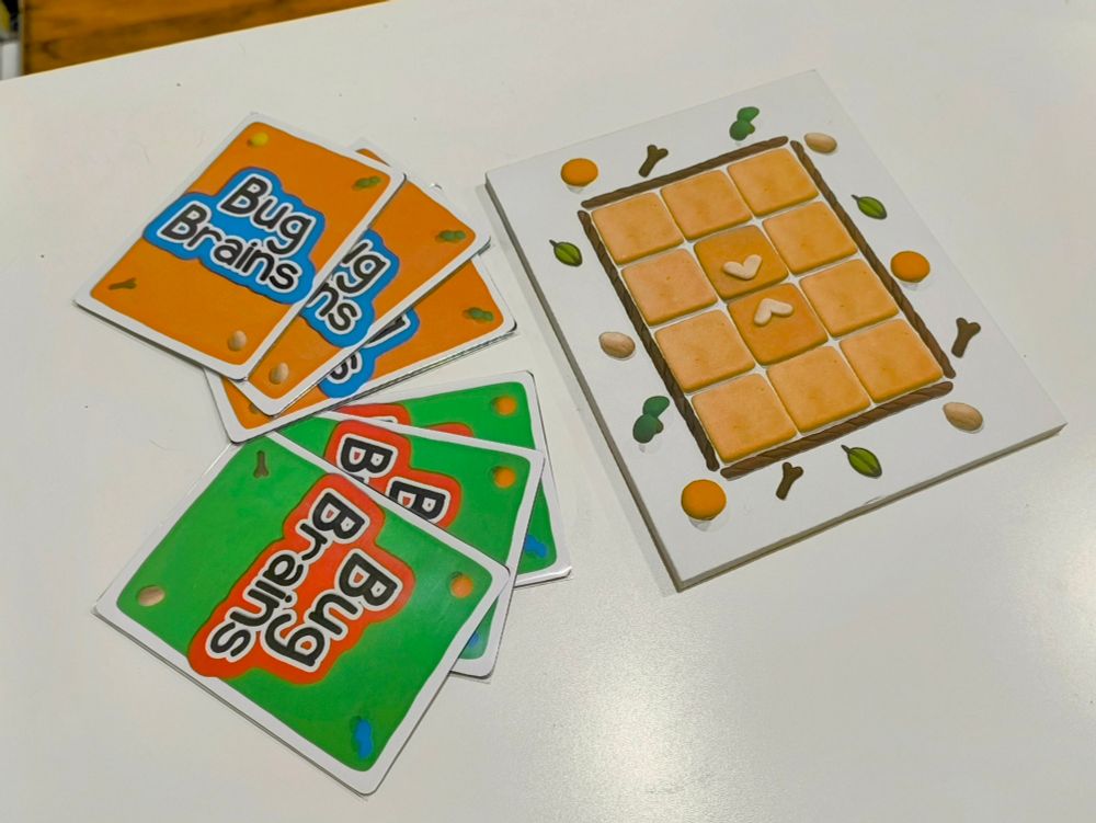 Two sets of three face down cards next to a 3 by 4 grid grid board, the cards say 'Bug Brains' on the backs. One set is orange and blue, the other is green and red. 
The tiles making up the grid on the board are a sandy brown. There are little decorative twigs and leaves and stuff around the borders of both the cards and the board. All the art was made with playdough.