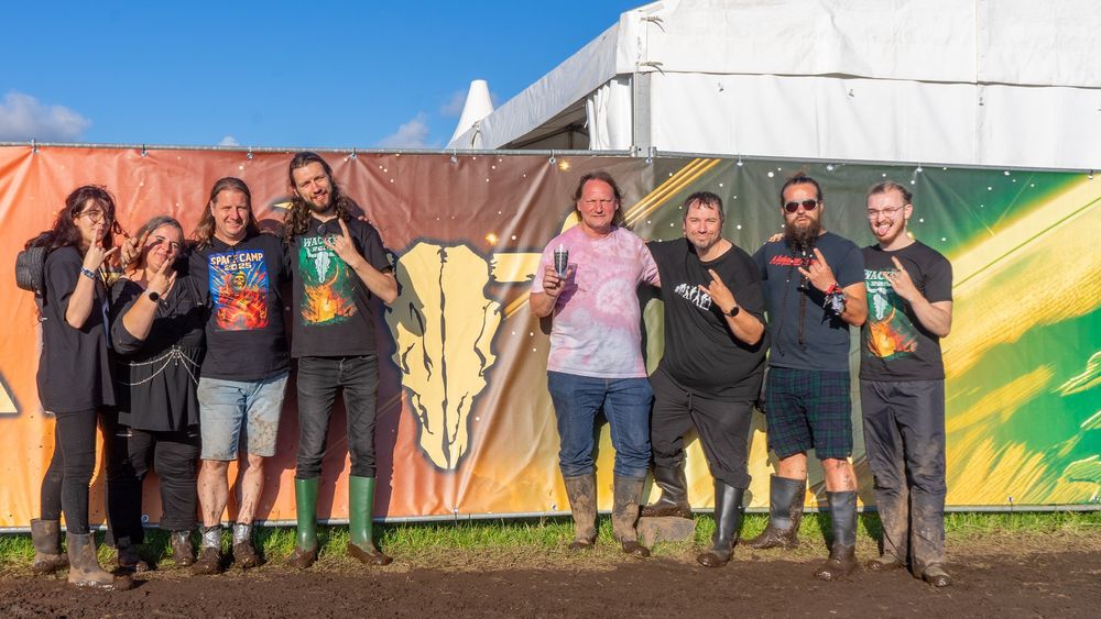 Das Space-Camp-Team in dreckigen Gummistiefeln und mit Metalhead-Pose vor einer Absperrwand mit Wacken-Logo. Davor der legendäre Schlamm