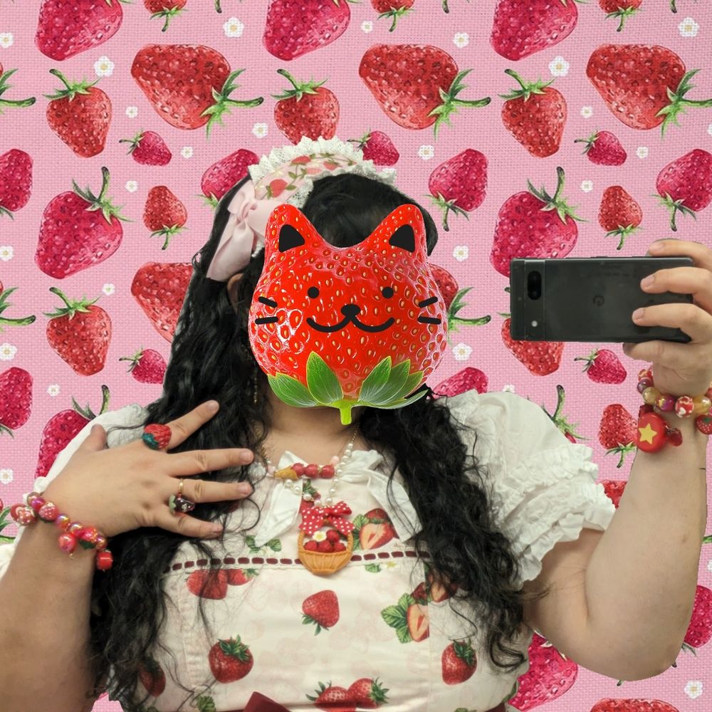 A mirror selfie of Poster onto the same pink strawberry background; while they have a face, a cat strawberry sticker is covering it. Here the embroidery of pinkish-red strawberries on their headdress is more visible. They have two strawberry rings on one hand, and a red strawberry charm bracelet on each wrist. The ring-less hand also has a tamagotchi charm on the bracelet. For necklaces, they have a basket of strawberries necklace, and a necklace of strawberries in an ice cream cone. Their super cute strawberry earrings with dangling flowers are not that visible in any of the pictures, but they're there!