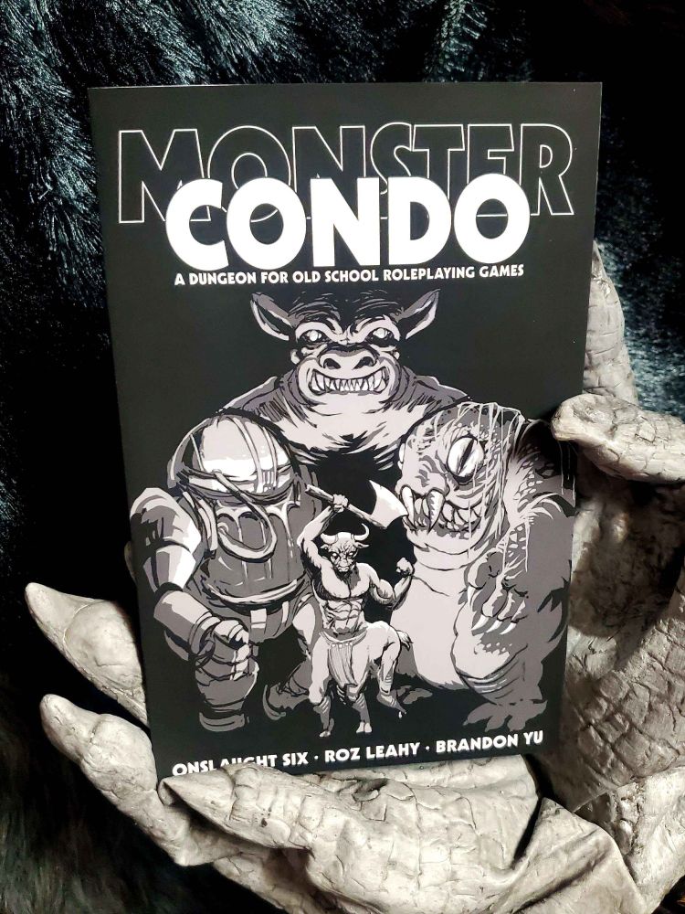 A furry body and gloved skeletal hand holding a zine-sized RPG book titled "MONSTER CONDO," featuring a demon, robot, swamp creature and goat centaur. 