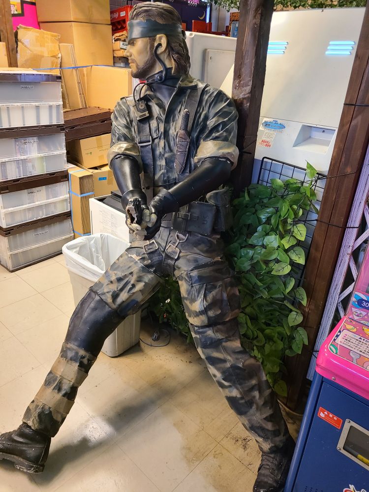 Life-size model of Solid Snake in MGS3 camouflage style, leaning stealthily at the corner of some intersecting aisles, peering expectantly with gun drawn