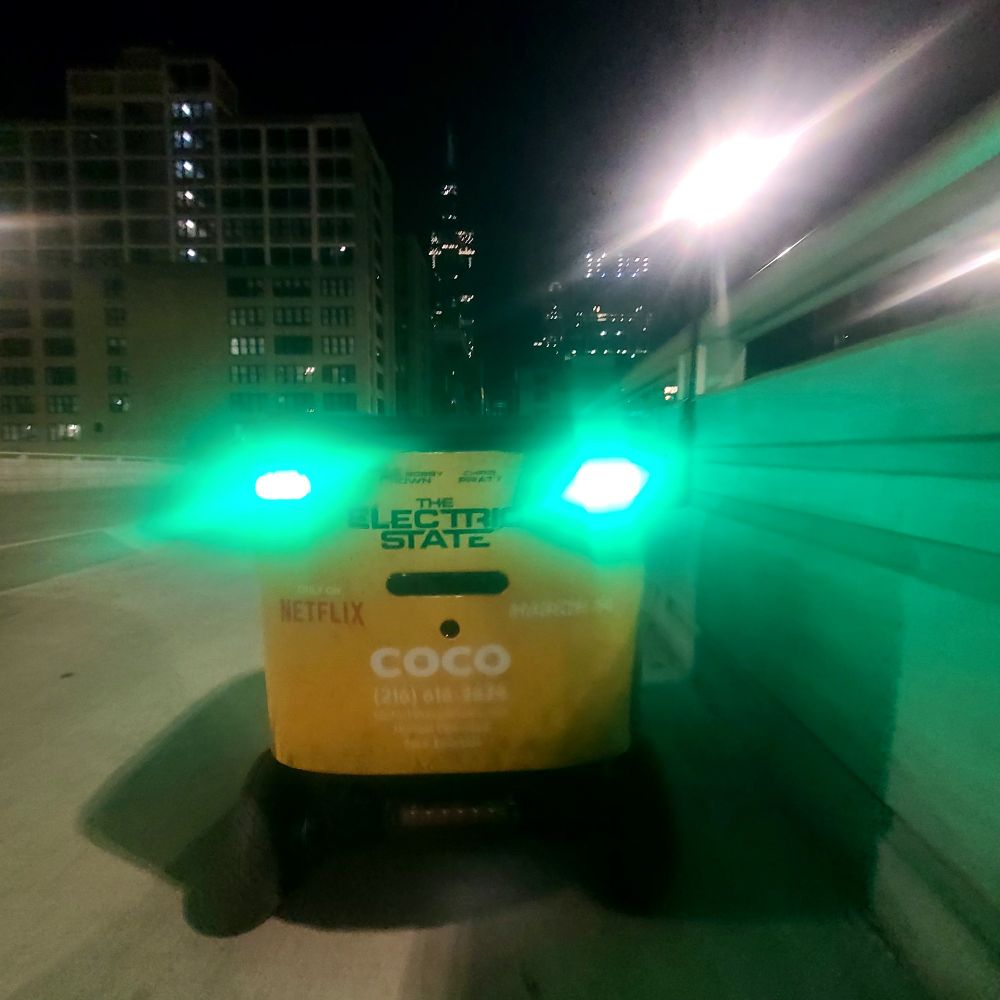 A yellow and black delivery robot stranded on a bridge in downtown Chicago, skyscrapers in the background, nighttime. 