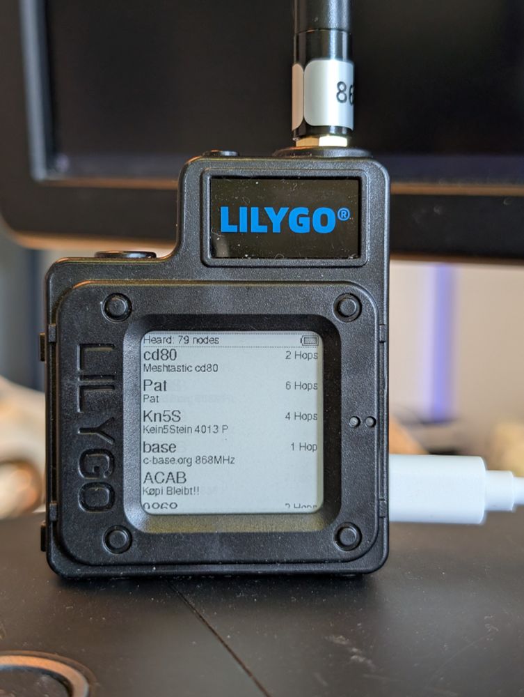 Photo of a LILYGO T-Echo resting on a workstation computer. The display lists recently seen Meshtastic nodes, among others the famous C-Base space station or the Køpi squat in Berlin, exclaiming "ACAB."

