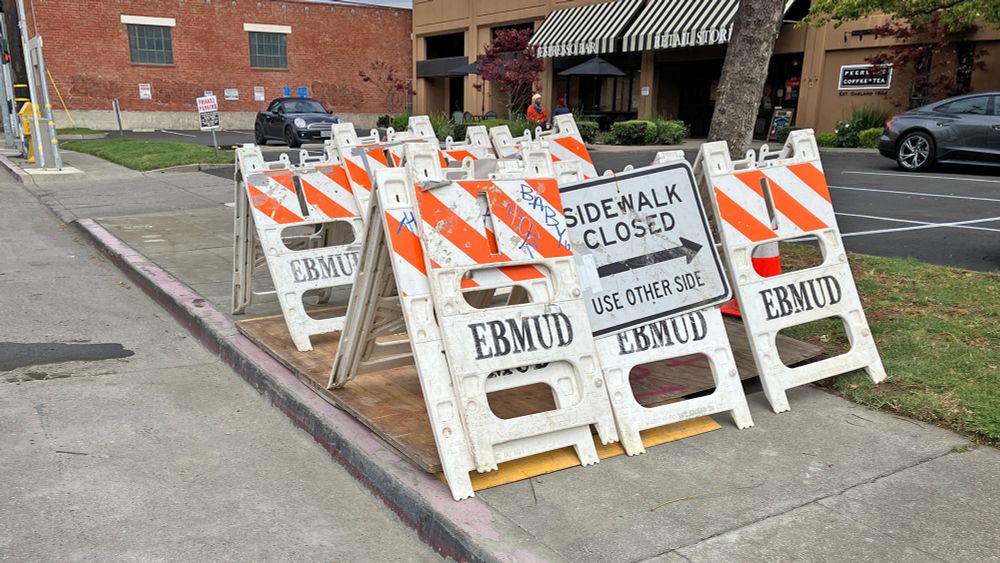 Set of portable orange-striped construction barriers labeled EBMUD with a sign on them says "sidewalk closed use other side"