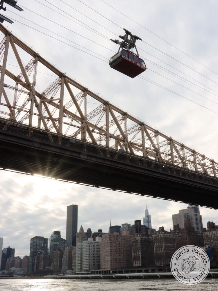 The Roosevelt Island Tramway is passing over the East River alongside the Queensboro Bridge in New York City. Upper East Manhattan is shown in the background.