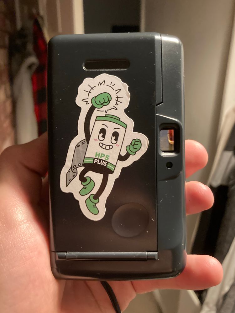 In a white man's hand is a film point and shoot camera, held portrait, with a sticker of anthropomorphic black, green, and white film canister stuck just beneath the viewfinder at the right of the camera, and beneath the film window at the top of the film door.