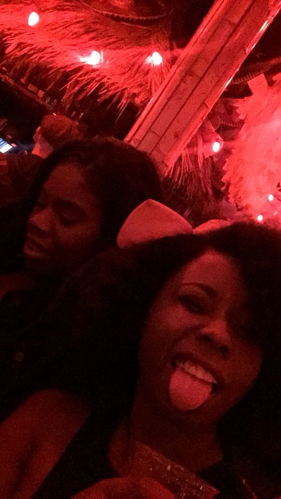 Photo of me in club with red lighting sticking out my tongue, eyes squint bow in my curly Afro hair with a drink in my hand