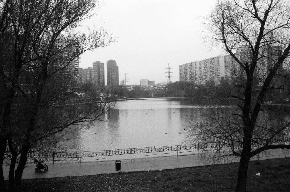 This monochrome photograph shows a pond in southern Moscow. Numerous high-rise buildings are visible on the left and right sides of the image, and there are a couple of trees close to the vantage point. A paved walkway runs alongside the pond and is separated from it by a short fence. A trash bin and a parked stroller are on the walkway. The photo was taken in the afternoon on a cloudy day.