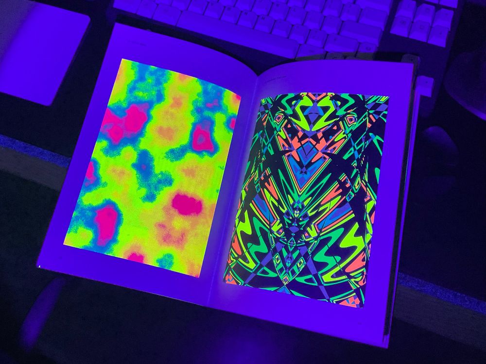 Imagen de una revista iluminada con luz negra. Muestra varias ilustraciones abstractas impresas en colores vivos fluorescentes que destacan enormemente sobre el fondo: un escritorio con varios objetos como un teclado, un ratón, etc.