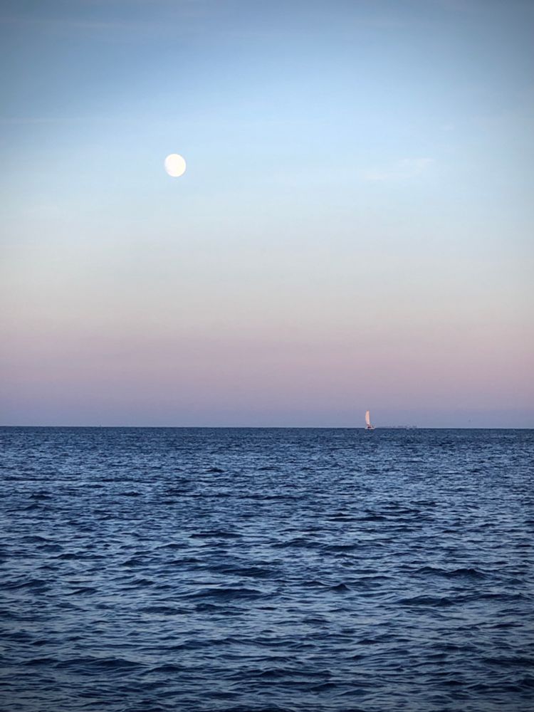 Blick auf den beginnenden Sonnenuntergang über dem Meer. Der Himmel verschwimmt in rosa und blau. Der Mond steht am Himmel und in der Ferne sieht man die Segel eines Bootes.
