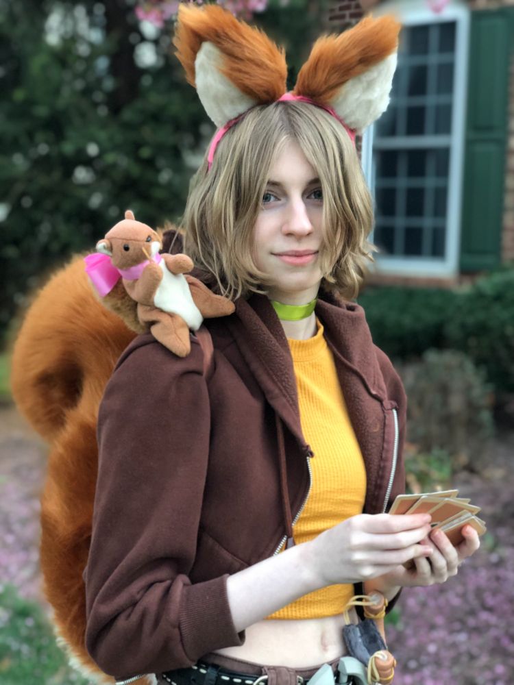 My daughter wearing her Squirrel Girl costume - fuzzy ears, brown jacket, Best Squirrel Friend Tippy Toe (plushy with a pink bow) on her shoulder, yellow shirt, standing in front of our home.  She's holding a bunch of "Deadpool's Guide to Supervillains" trading cards in her hand.