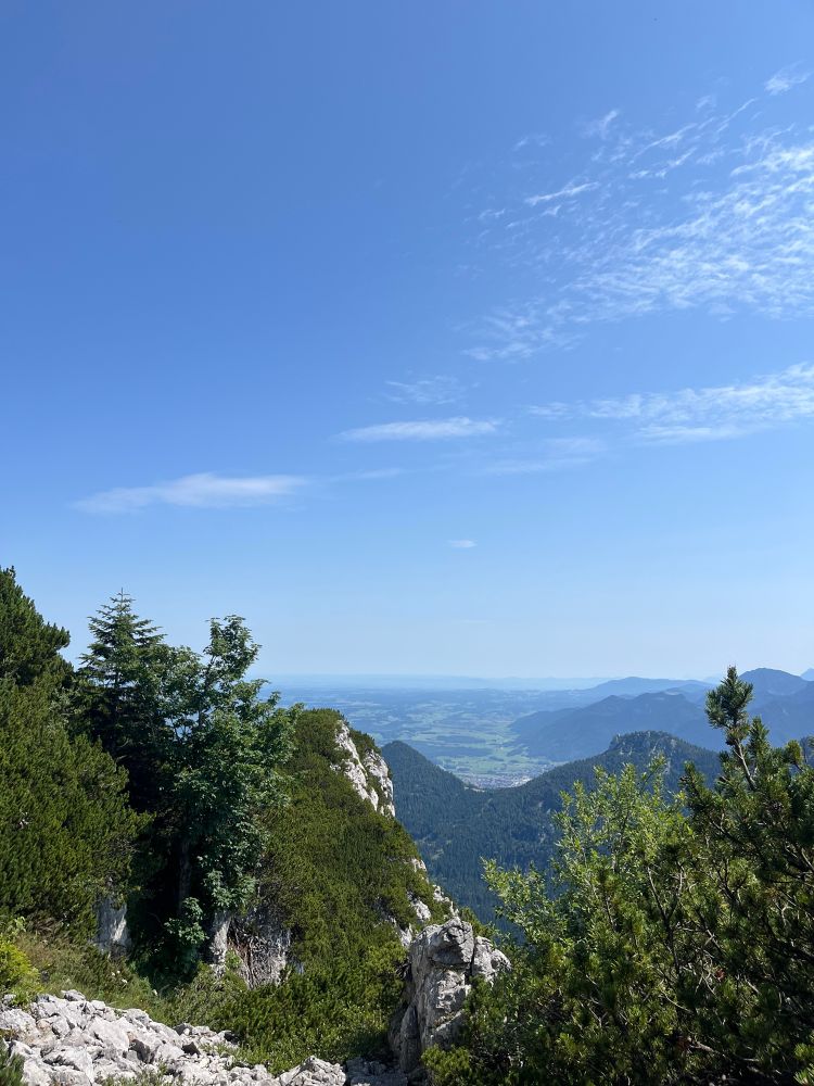 Blick vom Wanderweg kurz vor dem Ostgipfel der Kampenwand auf eine felsige Bergkante mit grünen Bäumen und Sträuchern im Vordergrund. Im Hintergrund weite Aussicht über bewaldete Hügel und Täler bis hin zur flachen Landschaft, unter einem klaren blauen Himmel mit wenigen, feinen Wolken.