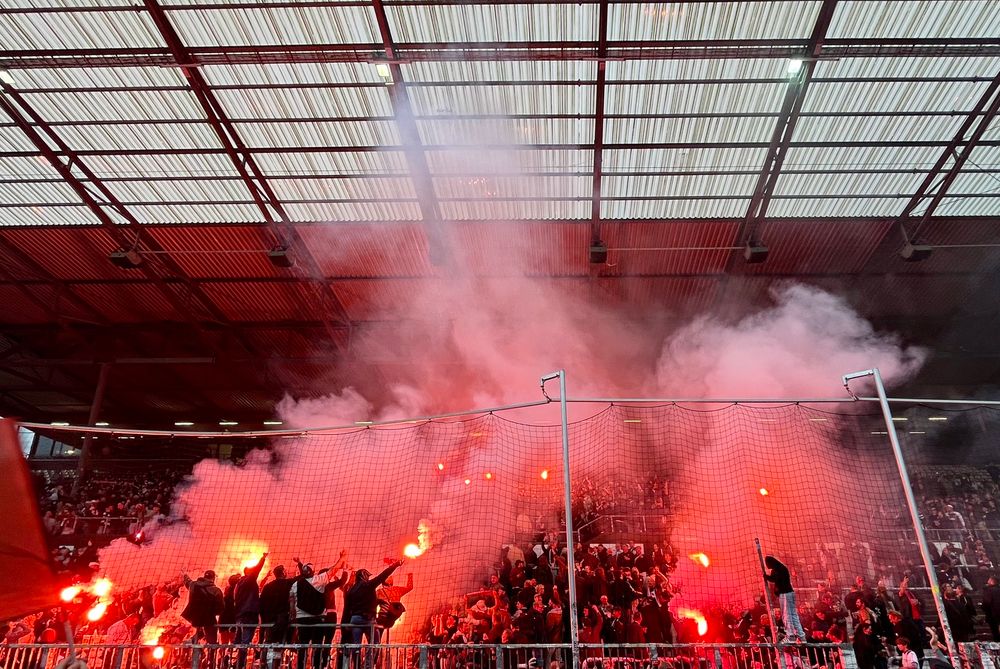 Im Millerntorstadion in der Südkurve stehen zahlreiche Fans dicht gedrängt im Block hinter einem Fangnetz. Viele von ihnen halten bengalische Feuer in den Händen, deren rotes Licht die Szene dramatisch beleuchtet. Dicker Rauch steigt auf und füllt das Stadiondach, das aus einem Gitter aus Metallträgern und transparenten Platten besteht. Die Stimmung wirkt leidenschaftlich und hitzig.