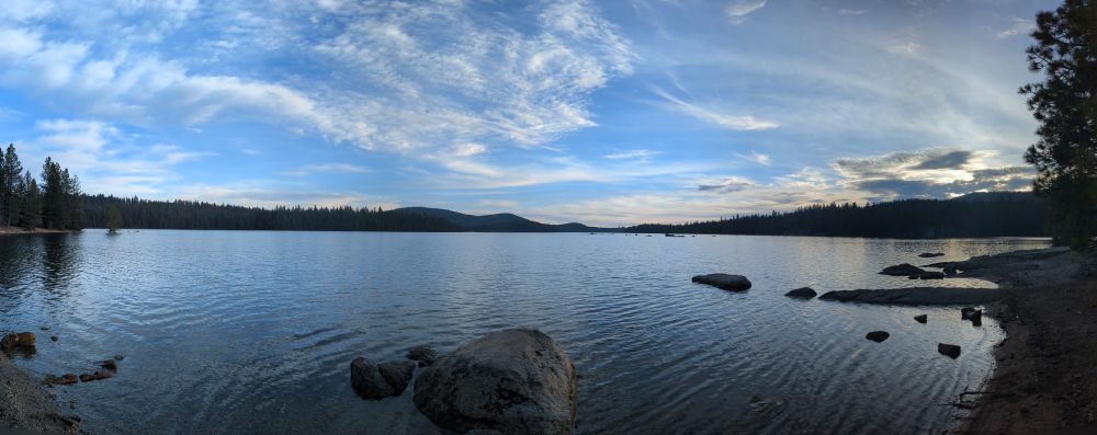 Photo of a calm lake near sunset. Hills with trees in background. Blue sky with light wispy clouds.