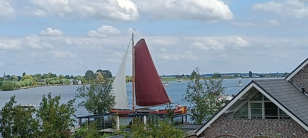 A tall ship with a dark red mainsail and a white headsail (jib?) on a lake with a house, trees and garden in the foreground. A light blue sky and fluffy white clouds behind.