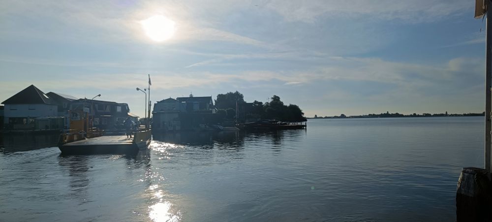 Small ferry to the left with a view of the lake opening to the right. Bright sun overhead.