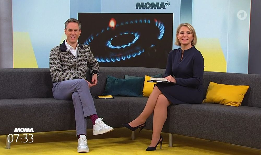Screenshot aus dem Studio des ARD Morgenmagazins (MOMA).
Zwei Moderatoren sitzen auf einem grauen Sofa mit gelben Kissen und schauen dabei in die Kamera.
Links Sven Lorig, rechts Susanne Link.
Im Hintergrund die Einblendung eines Gasherd-Kochfeldes.