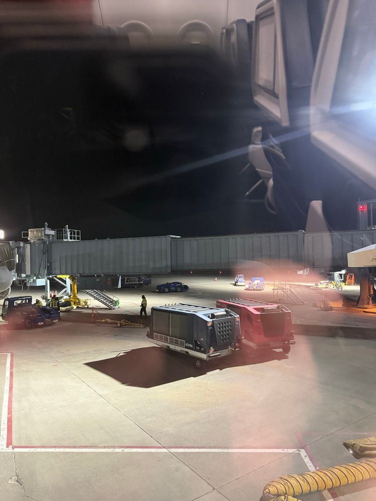 A picture taken from inside an airplane out the airplane window. The plane is on the ground, and the picture shows the jet bridge for the next gate with some workers outside. It’s still dark outside.