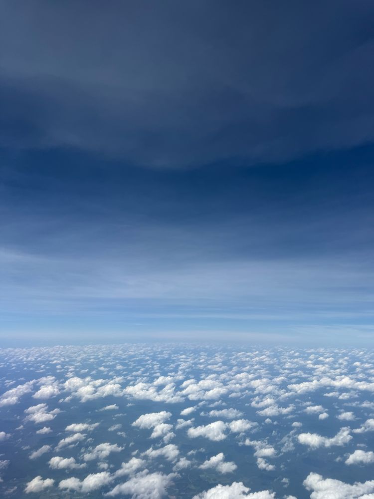 A photo taken from an airplane of the. There are small white fluffy clouds scattered below and a slightly hazy blue sky with white wisps above them. 