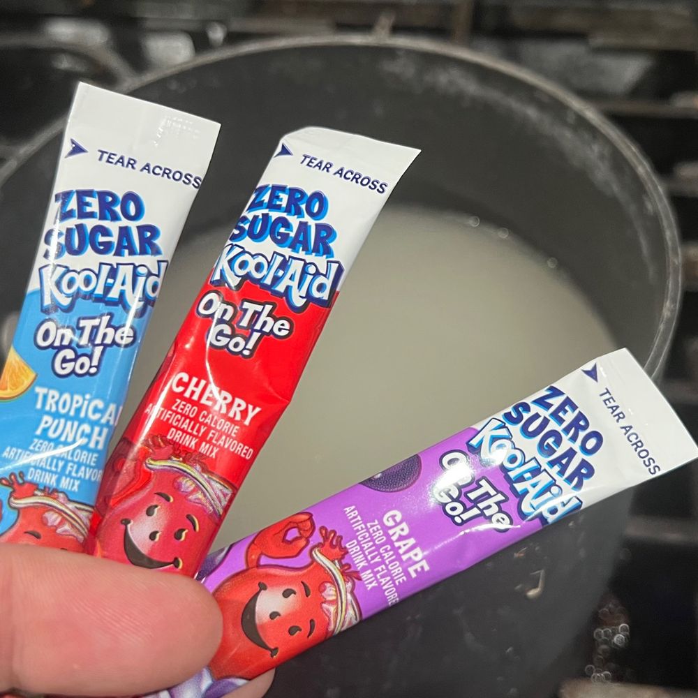 Three individual serving-size packets of Kool-Aid powder in the foreground, held in hand. The flavors are Tropical Punch (blue), Cherry (red), and Grape (purple). In the background behind them, a small pot on a stove containing a boiling, whitish fluid. This is equal parts sugar and water mixing until dissolved to make the simple sugar.