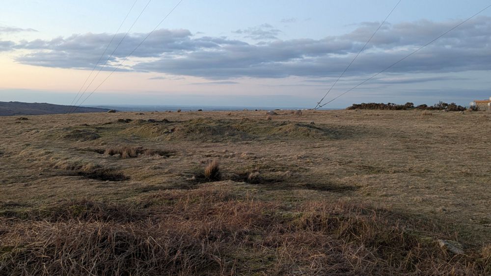 Colour photo of a grassy landscape. The sky is pale blue and slightly orange, on the ground the shadows show the rings of a cairn.