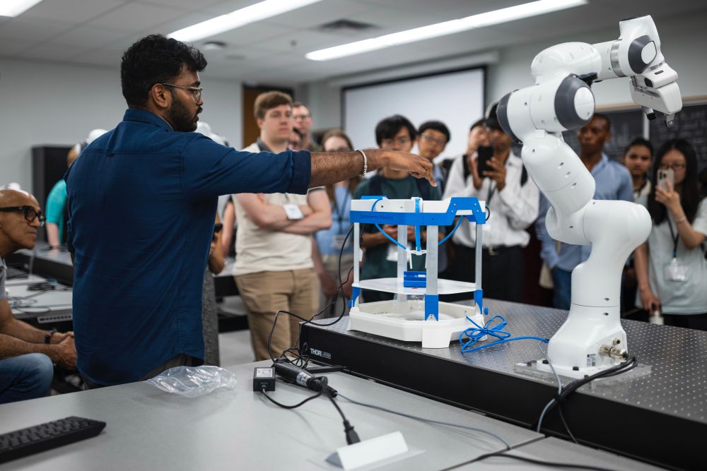 An individual demonstrating a robotic arm to a group of attentive onlookers in a high-tech workshop setting.
