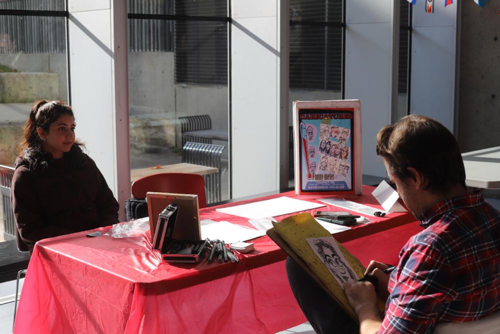 Two people sitting at a table outdoors, with one drawing a caricature for the other. 
