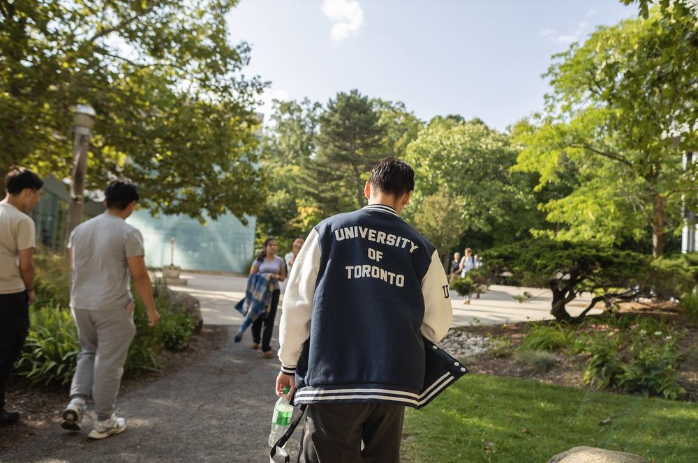 A person wearing a 'University of Toronto' jacket walking along a path in a campus setting with trees and other pedestrians nearby.

