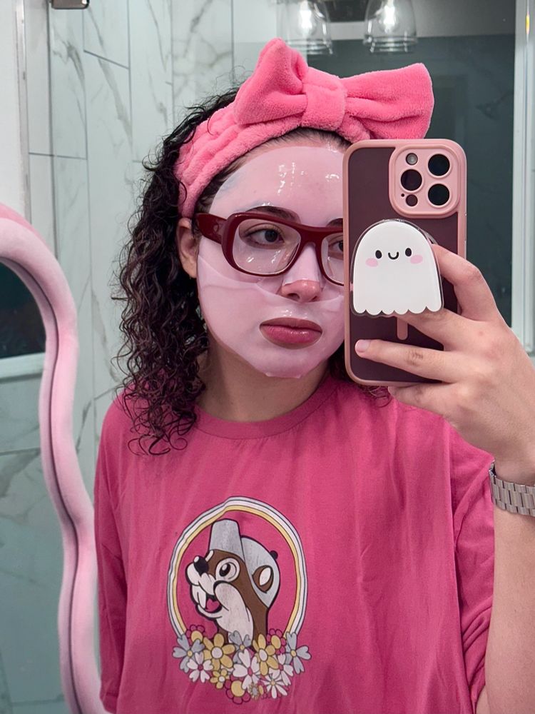 A person is taking a mirror selfie in a bathroom while wearing a pink sheet mask, red glasses, and a large pink headband with a bow. They are dressed in a pink oversized T-shirt with a cartoon animal surrounded by flowers and a rainbow on the front. The phone case features a cute ghost pop socket. The person has curly hair peeking out from the headband, and a watch is visible on their wrist. The background includes a marble shower wall and a pink-framed mirror.