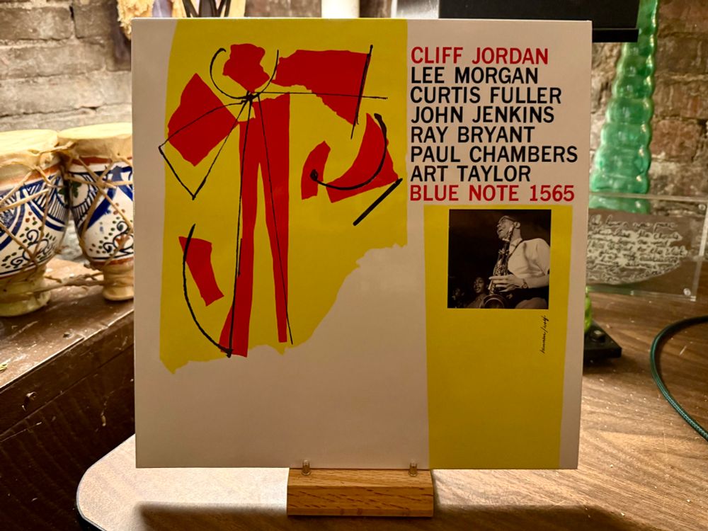 The photo shows a vinyl record displayed upright on a small wooden stand. The album cover is dominated by a modern abstract design with red and black shapes on a yellow background to the left, and a clean white section to the right. On the right side, text in bold capital letters lists the artists: 

CLIFF JORDAN, LEE MORGAN, CURTIS FULLER, JOHN JENKINS, RAY BRYANT, PAUL CHAMBERS, ART TAYLOR with BLUE NOTE 1565 in red and blue. 

Below the text is a small black-and-white photo of a saxophonist performing. The setting appears to be a warm, rustic indoor space with ceramic mugs, a brick wall, and decorative objects in the background.